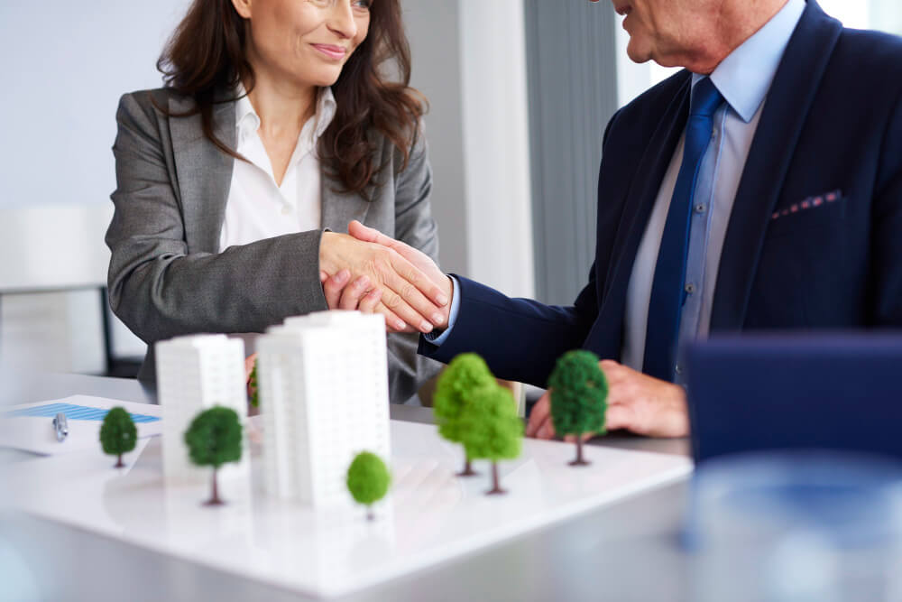 Two business professionals shaking hands over a model of buildings and trees on a table.