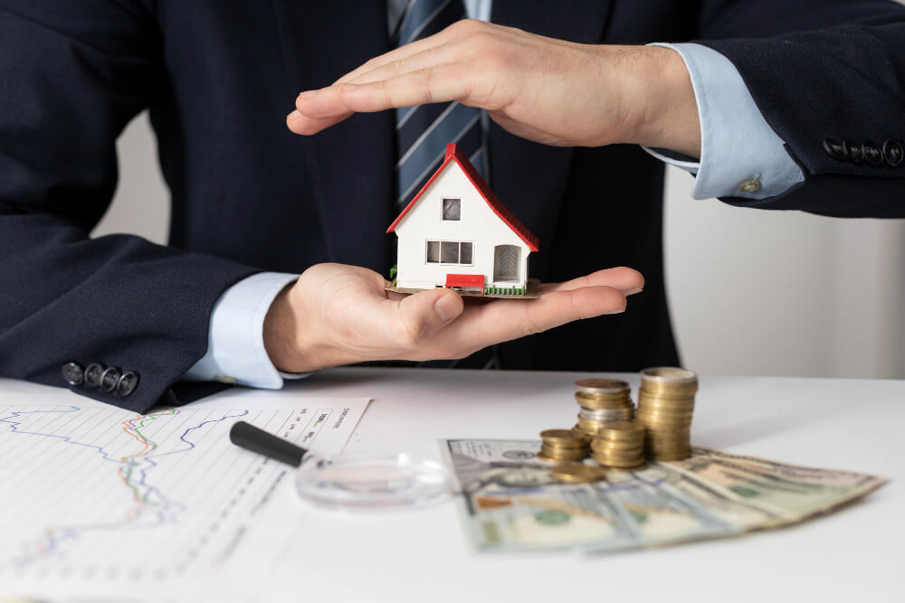 Person holding a small house model above a table with charts, coins, and dollar bills.