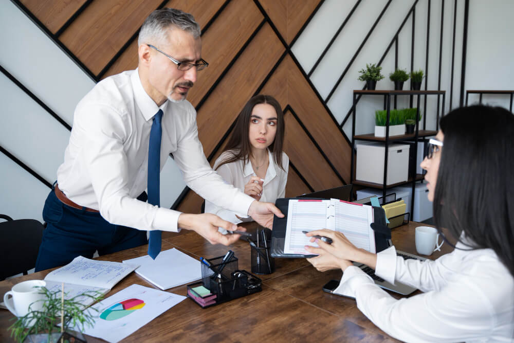 Office meeting with three people discussing documents at a table with charts and papers.